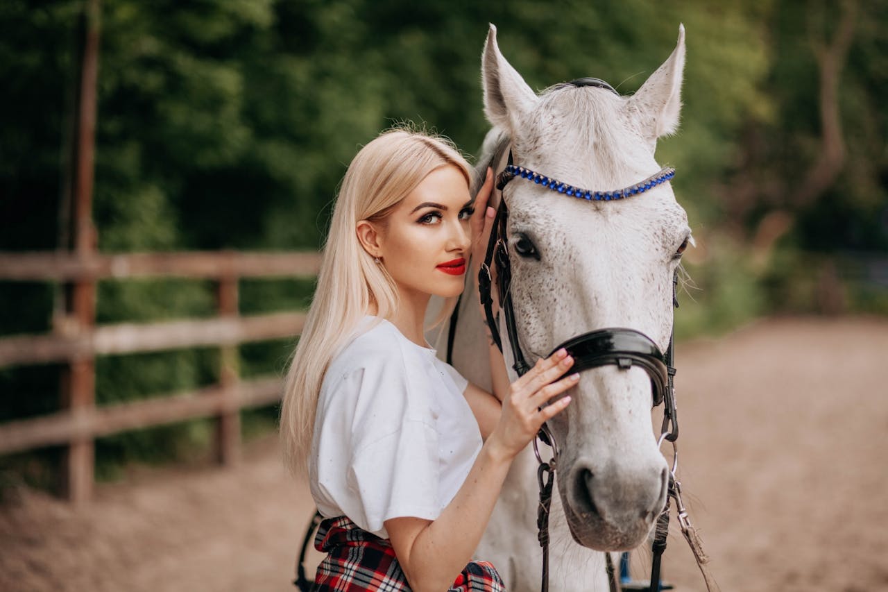 Blonde woman stands with white horse outdoors on bright day.
