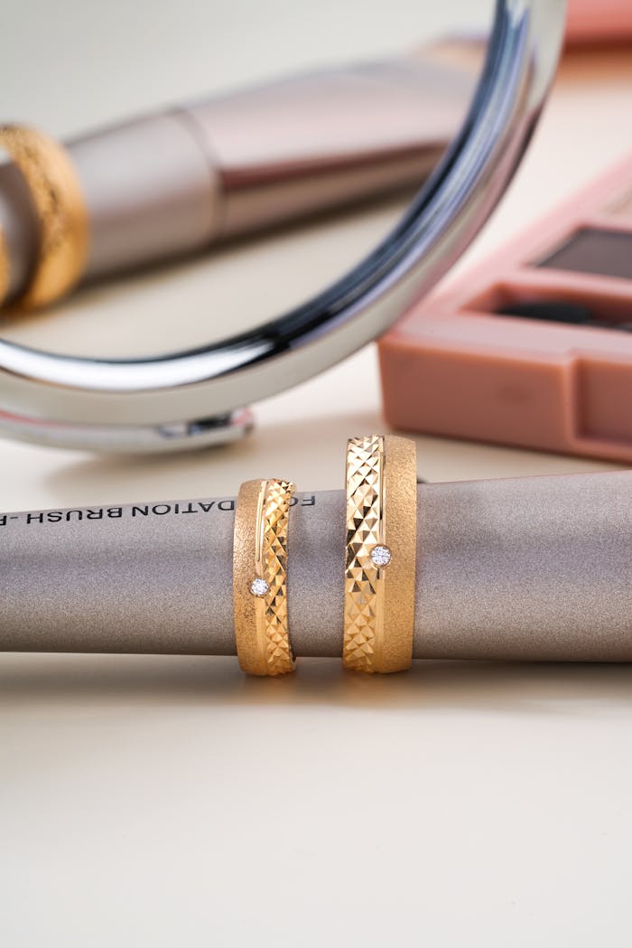 gallery-2 Stylized photo showcasing elegant gold wedding rings with diamond accents on a makeup brush.